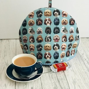 May include: A blue tea cosy with a pattern of many different dogs' faces. A cup of tea and a Lotus biscuit are on a white wooden table.