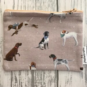 Può includere: Una pochette beige con cerniera con un motivo ripetitivo di cani marroni, neri e bianchi. La pochette ha un'etichetta bianca con il testo "jtaylorcrafts" stampato su di essa.