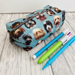 May include: A blue pencil case with a pattern of dog faces. The case is lying on a white wooden surface with three pens next to it.