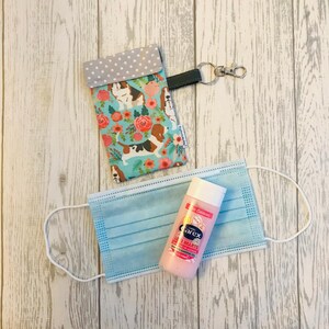 May include: A blue and white fabric pouch with a floral and dog print, a blue surgical mask, and a pink bottle of hand sanitiser with the text "Carex Gentle Hearts" on the label.
