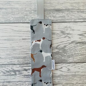 May include: A grey fabric bookmark with a white ribbon loop and a repeating pattern of brown and white dogs. The bookmark has a white label with the text "jtaylorcrafts" printed on it.
