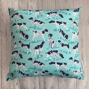 May include: Square throw pillow with a teal background featuring a repeating pattern of black and white spotted dogs in various poses. The pillow has a soft, textured appearance, suggesting a comfortable fabric.