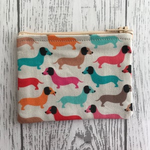 Peut inclure: Une petite pochette zippée avec un fond blanc et un motif répété de chiens teckels colorés. Les chiens sont de différentes couleurs, notamment orange, rose, bleu et marron.