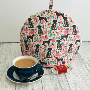 May include: A white tea cosy with a floral pattern featuring brown and black dogs. The cosy has a pink trim and a loop handle. A blue teacup with a saucer and a silver spoon are on a white wooden table. A packet of Lotus biscuits is on the table.