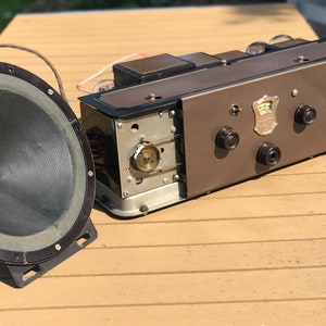 May include: Vintage radio with a brown and black exterior and a speaker. The radio has a metal grill and two knobs. The speaker is black and has a round shape.