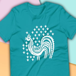 Puede incluir: Una camiseta azul turquesa con un diseño de gallo blanco. El gallo está rodeado de un círculo de estrellas blancas.