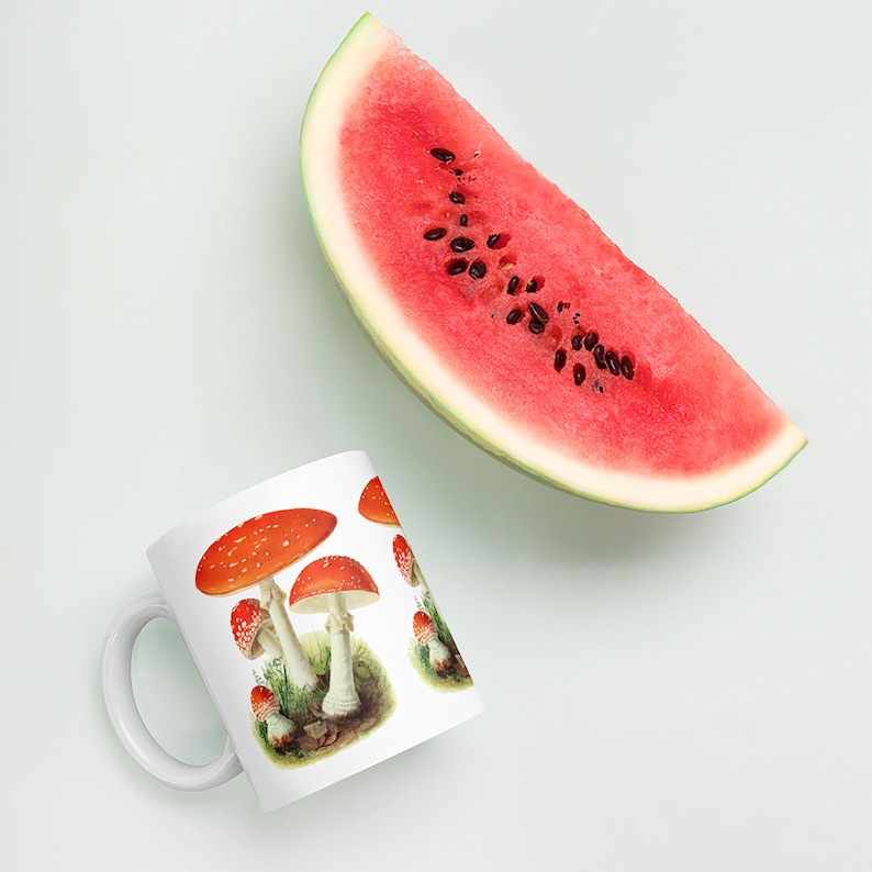 K&ouml;nnte beinhalten: Wei&szlig;e Keramiktasse mit einem sich wiederholenden Muster aus rot-wei&szlig; gepunkteten Pilzen. Eine gro&szlig;e Scheibe Wassermelone mit schwarzen Samen befindet sich im Hintergrund. Die Tasse hat einen wei&szlig;en Henkel.