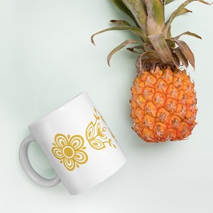 Pu&ograve; includere: Una tazza in ceramica bianca con un motivo floreale dorato. La tazza &egrave; accanto a un ananas intero su uno sfondo verde chiaro.