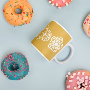 Pu&ograve; includere: Una tazza gialla con un motivo floreale bianco. La tazza &egrave; circondata da ciambelle con codette colorate e glassa rosa.