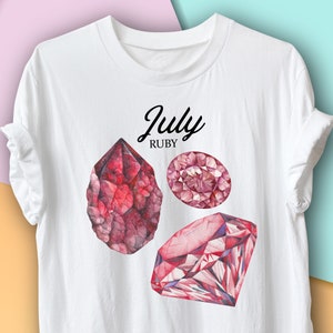 Può includere: T-shirt bianca con un disegno ad acquerello di tre gemme di rubino. La maglietta ha il testo "July Ruby" in corsivo nero. Le gemme sono di varie forme e sfumature di rosso.