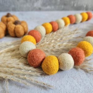 May include: A decorative felt ball garland in autumnal shades of orange, yellow, and cream. The garland is arranged in a curved line, with small felt pumpkins in the background. The garland rests on dried grass.