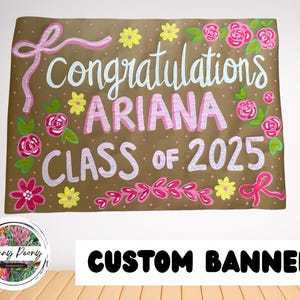 May include: A custom banner with the words "congratulations ARIANA CLASS OF 2025" in white and pink lettering. The banner is decorated with pink flowers, yellow flowers, and pink bows on a brown background.
