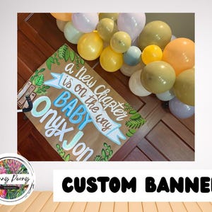 May include: A custom banner with the text "a new chapter is on the way BABY Onyx Jon" in blue and white lettering, with green leaf accents. The banner is surrounded by a colorful balloon arch.