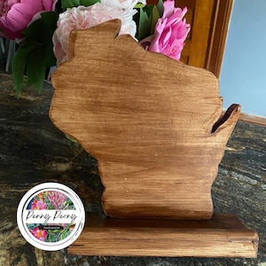 May include: A wooden cutout of the state of Wisconsin, stained in a warm brown hue. The state-shaped piece stands on a rectangular base. The Penny Peony logo is visible in the lower left corner. Pink and white flowers are in the background.