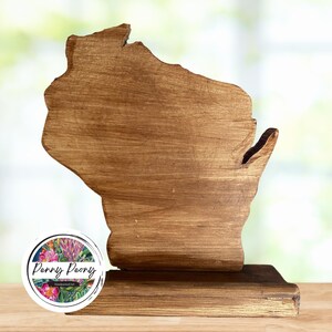 May include: A wooden cutout of the state of Wisconsin, stained in a warm brown hue. The state-shaped piece is mounted on a rectangular base. A circular logo with floral designs and the text "Penny Peony Handpainted Art" is visible.