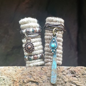 May include: Two beige and brown rope hair wraps with metal accents and turquoise beads. The hair wraps are on a brown textured surface.
