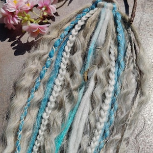 Op de afbeelding: Een set haar extensions met een mix van golvende lichtbruine, witte en blauwe strengen. De extensions zijn bevestigd aan een bruin leren hoofdband met een klein houten label waarop "Mizomoda" staat.
