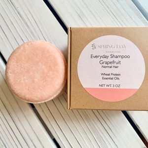 May include: A round, pink grapefruit shampoo bar in a cardboard box with a label that reads "SPRING DAY Everyday Shampoo Grapefruit Normal Hair Wheat Protein Essential Oils NET WT. 3 OZ"