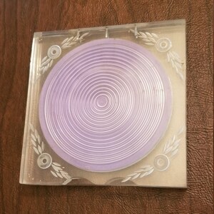 May include: A clear plastic square frame with a purple concentric circle design inside. The frame has a decorative design of leaves and circles around the edge.