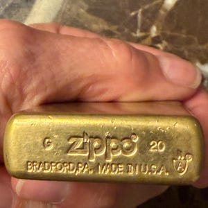 K&ouml;nnte beinhalten: Ein messingfarbenes Zippo-Feuerzeug mit rechteckigem Boden, auf dem der eingestanzte Text Zippo, 20 sowie BRADFORD, PA. MADE IN U.S.A. zusammen mit einem G- und A-Symbol zu sehen ist. Die Metalloberfl&auml;che ist goldfarben und weist Gebrauchsspuren auf.