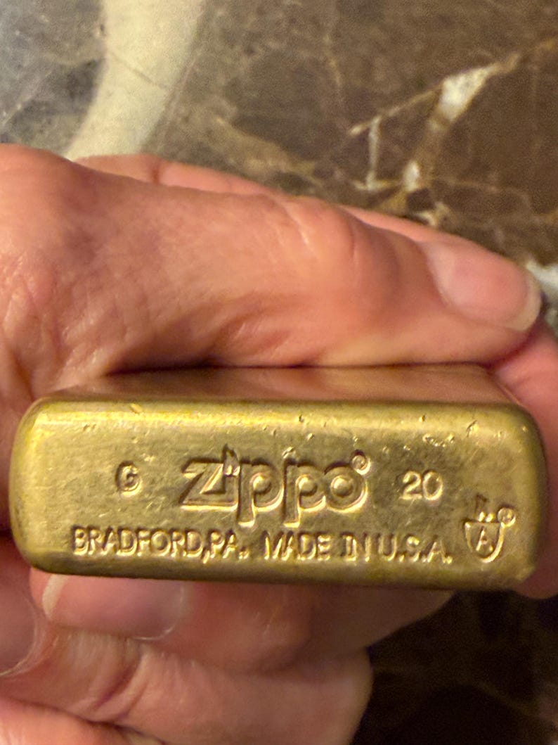 K&ouml;nnte beinhalten: Ein messingfarbenes Zippo-Feuerzeug mit rechteckigem Boden, auf dem der eingestanzte Text Zippo, 20 sowie BRADFORD, PA. MADE IN U.S.A. zusammen mit einem G- und A-Symbol zu sehen ist. Die Metalloberfl&auml;che ist goldfarben und weist Gebrauchsspuren auf.