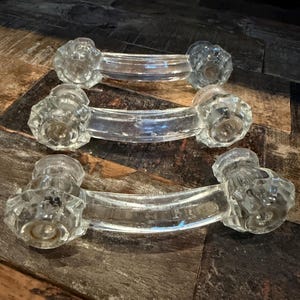 May include: Three clear glass cabinet pulls with faceted knob ends and curved handles. The pulls are arranged horizontally, showcasing their vintage design and potential for antique furniture restoration. Each pull is approximately 6 inches long.