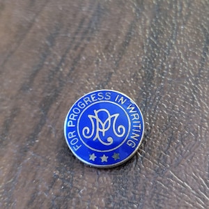May include: A round, blue pin with the text "FOR PROGRESS IN WRITING" in a white, circular border. The center features a stylized design and three stars. The pin is set against a brown surface.