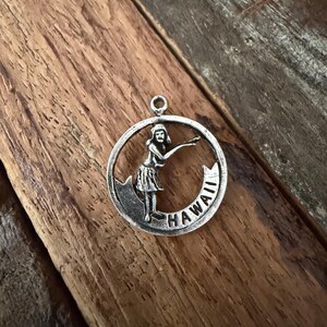 May include: A circular silver-toned metal charm featuring a raised relief of a hula dancer in a grass skirt. The dancer is positioned within the circular frame, which includes a banner at the bottom with the embossed text HAWAII.