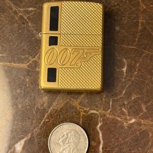 K&ouml;nnte beinhalten: Ein goldfarbenes Metallfeuerzeug mit einer texturierten Kreuzschraffur und schwarzen rechteckigen Akzenten. Auf der Vorderseite befindet sich das gepr&auml;gte 007-Logo. Eine US-Vierteldollarm&uuml;nze liegt unter dem Feuerzeug und dient als Gr&ouml;&szlig;envergleich.