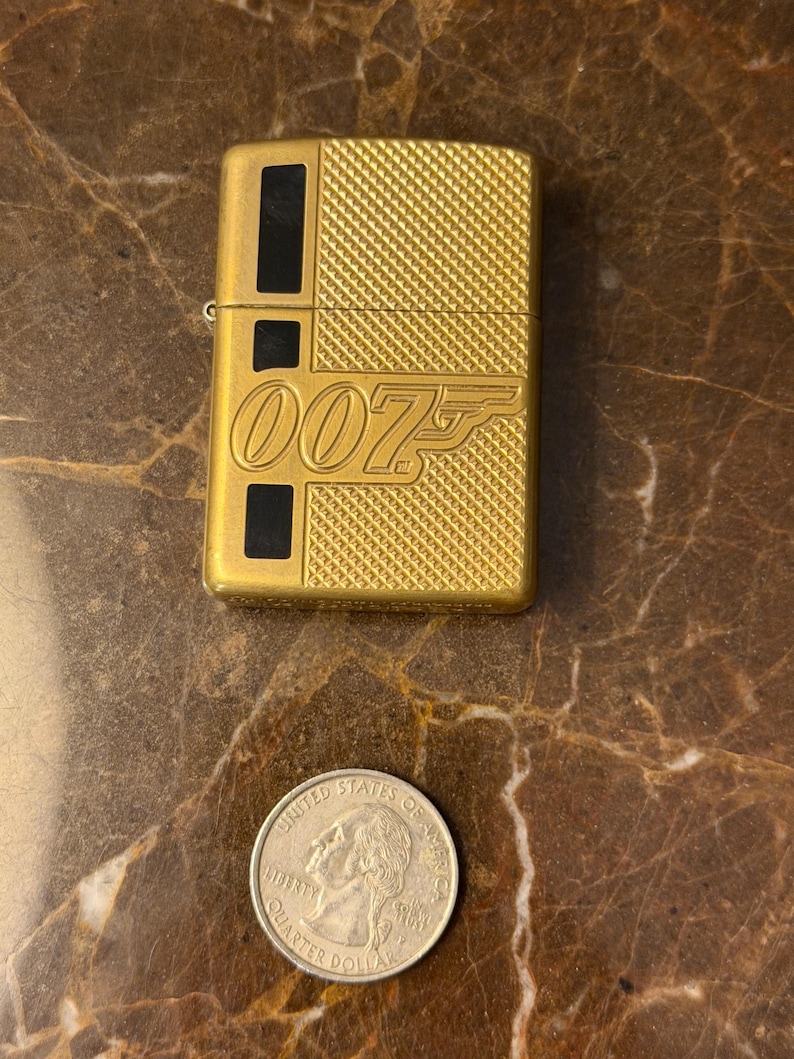 K&ouml;nnte beinhalten: Ein goldfarbenes Metallfeuerzeug mit einer texturierten Kreuzschraffur und schwarzen rechteckigen Akzenten. Auf der Vorderseite befindet sich das gepr&auml;gte 007-Logo. Eine US-Vierteldollarm&uuml;nze liegt unter dem Feuerzeug und dient als Gr&ouml;&szlig;envergleich.