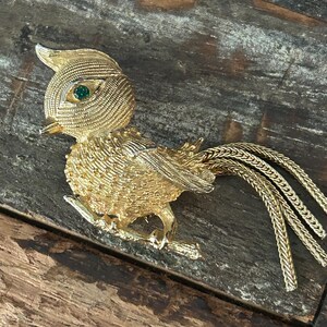 May include: A gold-tone bird brooch with green rhinestone eyes. The bird is perched on a branch and has a long, flowing tail.