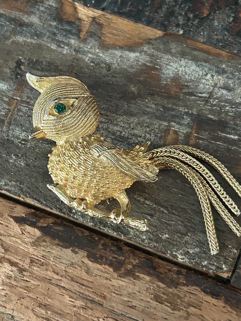 May include: A gold-tone bird brooch with green rhinestone eyes. The bird is perched on a branch and has a long, flowing tail.