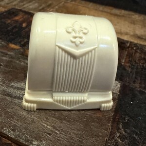 May include: A small, off-white, molded plastic trinket box with a rounded top. The front features a raised fleur-de-lis emblem centered above a vertical ribbed panel. The base has a stepped design with small, decorative feet at each corner.