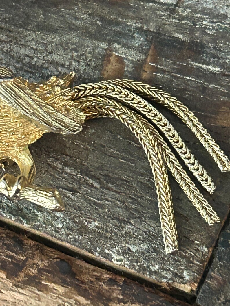 May include: Gold-toned metal brooch featuring a stylized bird with a long, braided tail.