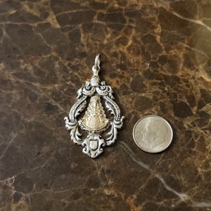 Op de afbeelding: Zilveren hanger met een goudkleurig beeld van de Maagd Maria. De hanger is versierd en draagt de woorden "Reconciliacion" en "Bendicion" rond het beeld. De hanger is ongeveer zo groot als een Nederlandse tien-cent munt.