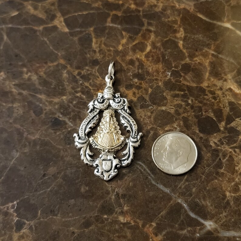 Op de afbeelding: Zilveren hanger met een goudkleurig beeld van de Maagd Maria. De hanger is versierd en draagt de woorden "Reconciliacion" en "Bendicion" rond het beeld. De hanger is ongeveer zo groot als een Nederlandse tien-cent munt.