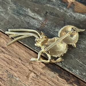 May include: Gold-toned brooch in the shape of a rooster with a long tail of gold chains. The brooch has a pin on the back.
