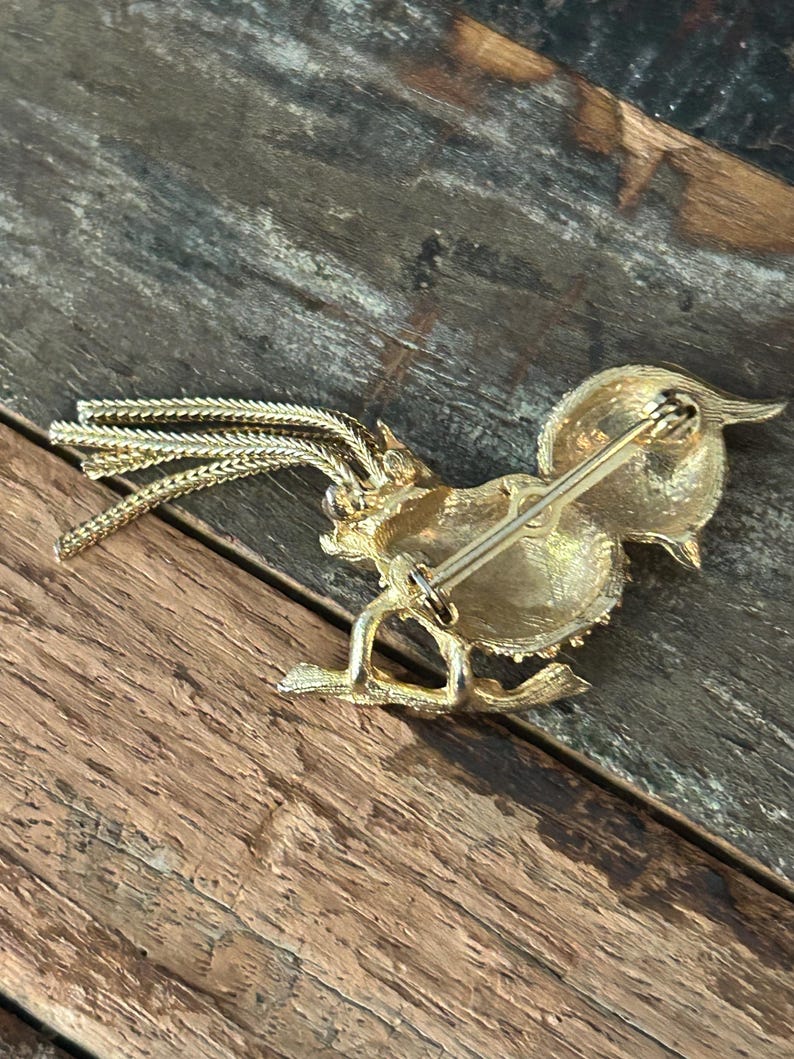 May include: Gold-toned brooch in the shape of a rooster with a long tail of gold chains. The brooch has a pin on the back.