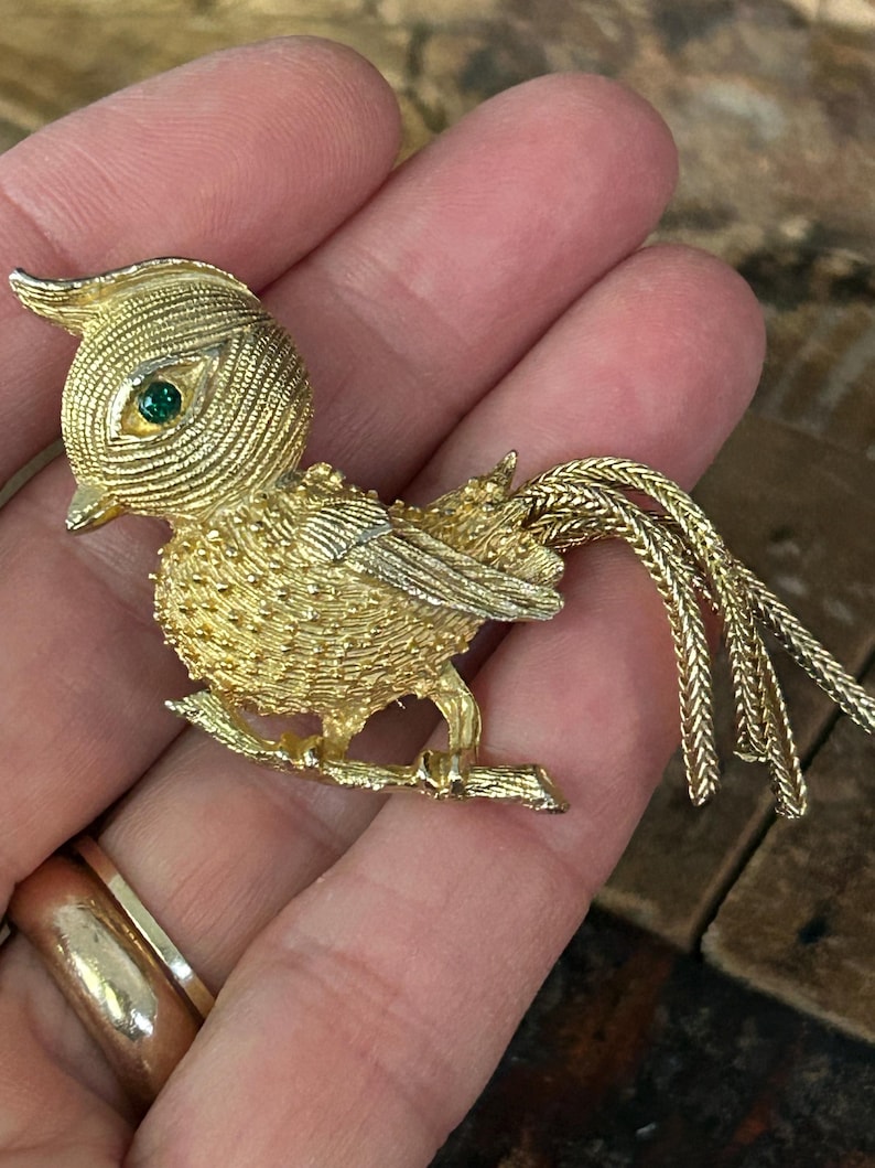 May include: A gold-tone bird brooch with a green rhinestone eye, perched on a twig with a long, braided tail.
