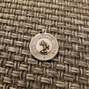 May include: A circular silver-toned metal charm featuring a raised relief of Mickey Mouse in the center. The text Walt Disney World is embossed around the perimeter of the charm, which has a textured border and a small loop at the top for attachment.