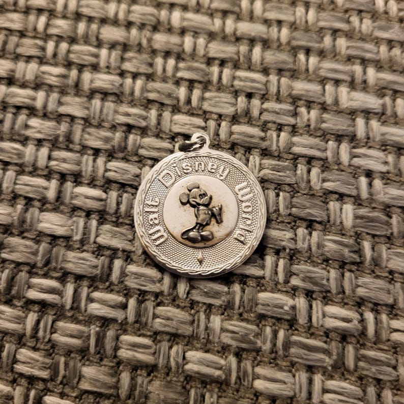 May include: A circular silver-toned metal charm featuring a raised relief of Mickey Mouse in the center. The text Walt Disney World is embossed around the perimeter of the charm, which has a textured border and a small loop at the top for attachment.