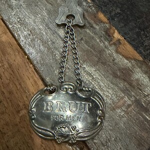 May include: A silver-toned metal bottle tag featuring a key-shaped top piece connected by two small chains to an oval-shaped plaque. The plaque is embossed with the text BRUT FOR MEN and features decorative scrollwork and a leaf motif at the bottom.