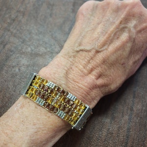 May include: A multi-strand bracelet featuring brown and gold beads with silver accents. The bracelet is made up of multiple rows of beads and has a silver clasp.