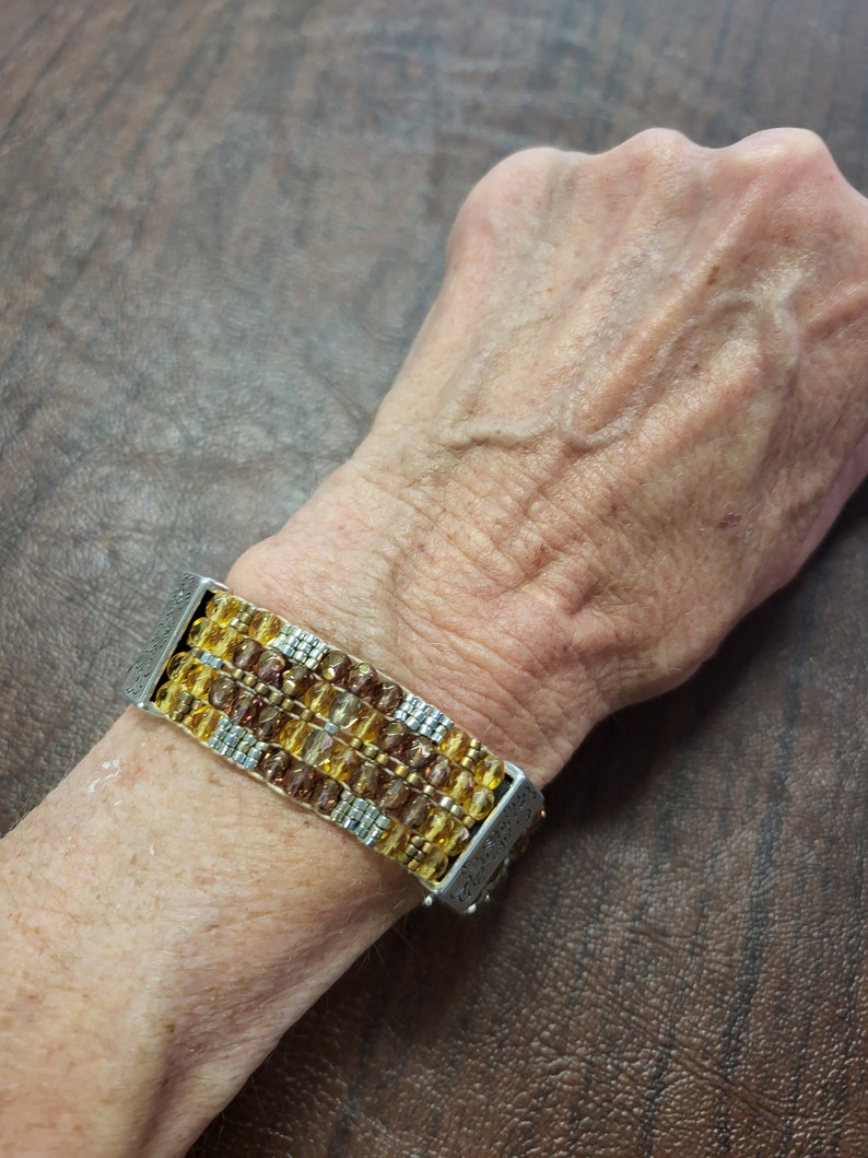 May include: A multi-strand bracelet featuring brown and gold beads with silver accents. The bracelet is made up of multiple rows of beads and has a silver clasp.