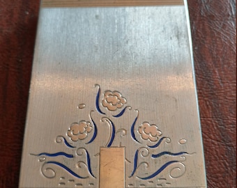Könnte beinhalten: Ein silbernes Kompakt mit einem blauen und goldenen Blumendesign. Das Kompakt hat einen Klappdeckel und einen kleinen Verschluss.