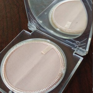 May include: A clear plastic compact with two round, white, fabric-lined compartments. The compact has a hinged lid with a mirror.