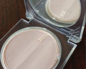 May include: A clear plastic compact with two round, white, fabric-lined compartments. The compact has a hinged lid with a mirror.