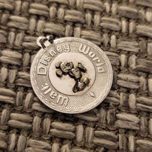 May include: A circular silver-toned metal charm featuring a raised relief of Mickey Mouse in the center. The text Walt Disney World is embossed around the perimeter. The charm has a textured border and a small loop at the top for attachment.