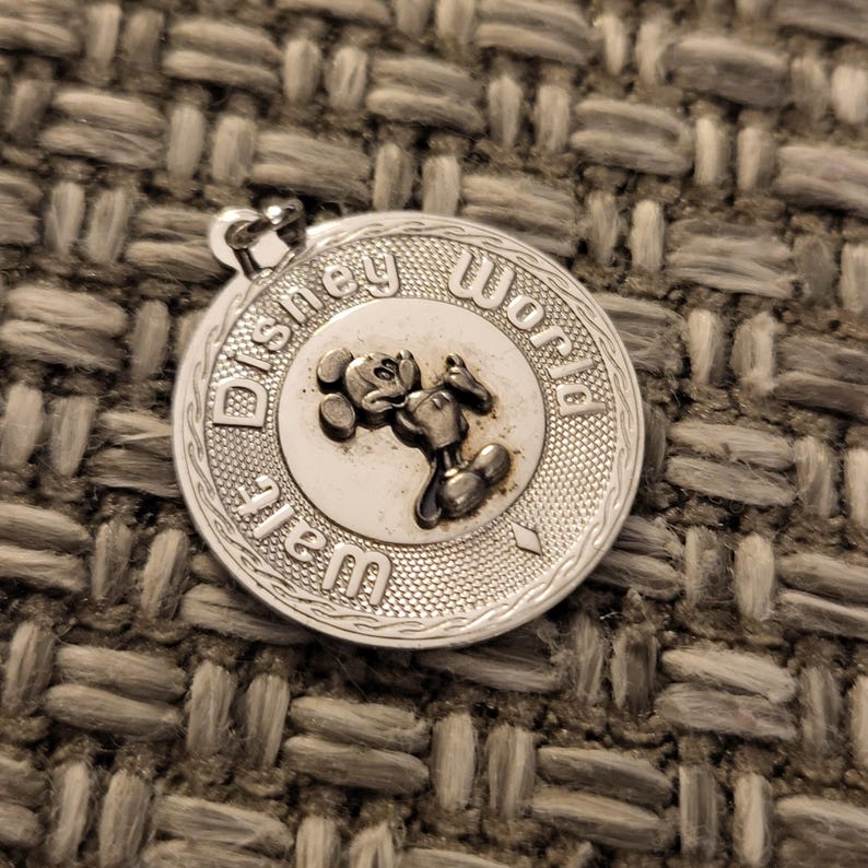 May include: A circular silver-toned metal charm featuring a raised relief of Mickey Mouse in the center. The text Walt Disney World is embossed around the perimeter. The charm has a textured border and a small loop at the top for attachment.