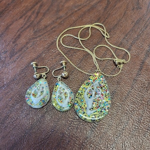 May include: A vintage jewelry set featuring a gold-tone necklace and matching clip-on earrings. The pendant and earrings have a teardrop shape with a white figure design, surrounded by colorful glitter and a gold wire mesh.
